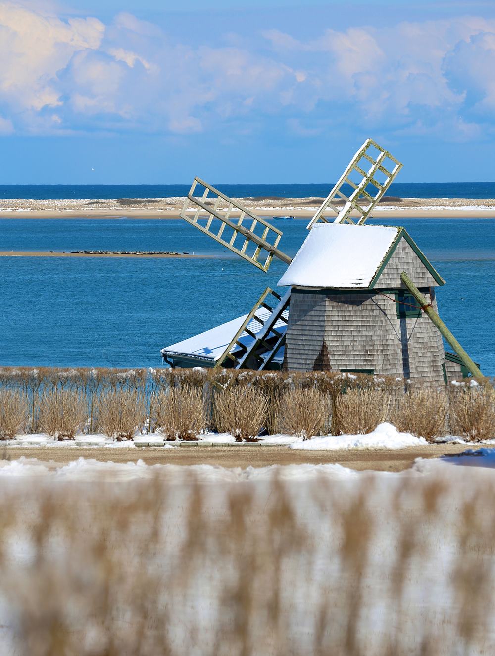 A shingled windmill under snow with the water behind it.