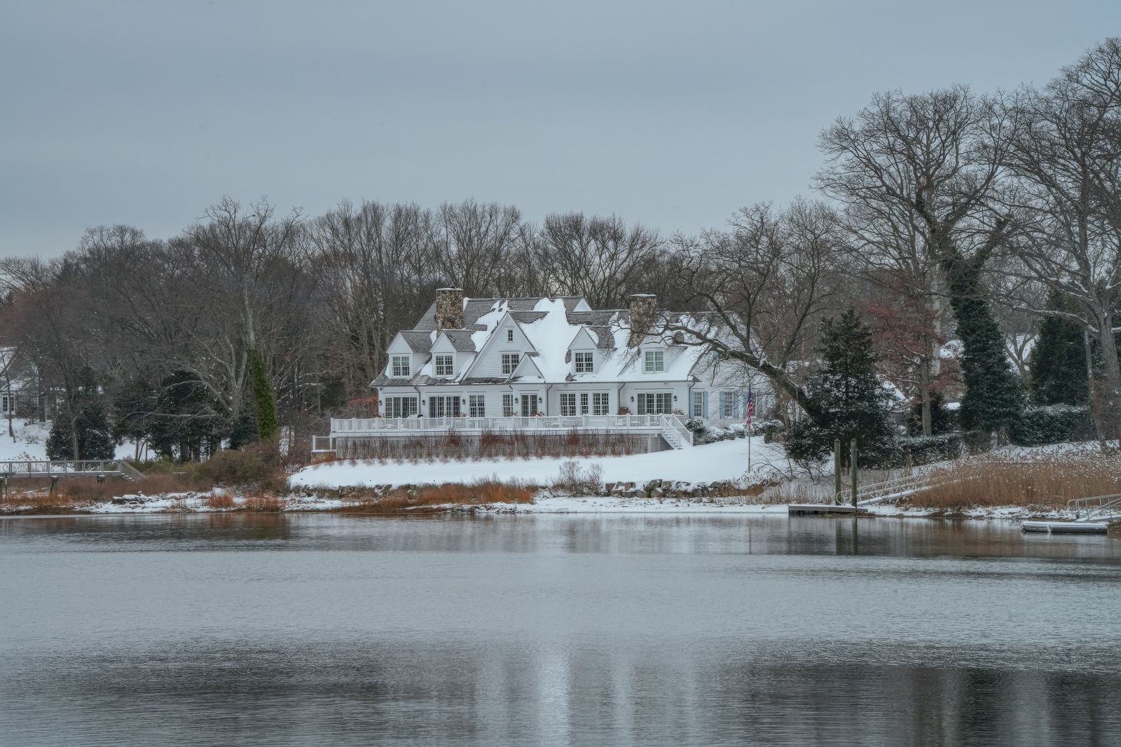 A large shingled Cape house standing closed up in the snow above the water.